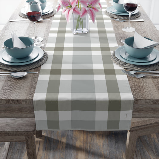 A rectangular table runner with a muted sage green and off-white buffalo plaid pattern, laid down the center of a wooden dining table set with blue bowls, plates, wine glasses, and pink lilies.