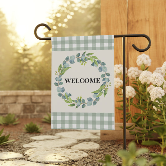 A decorative garden flag hanging from a black metal stand outdoors. The flag has a white center with a green and blue watercolor wreath and the word “WELCOME” in black text, plus pale green gingham bands at the top and bottom.